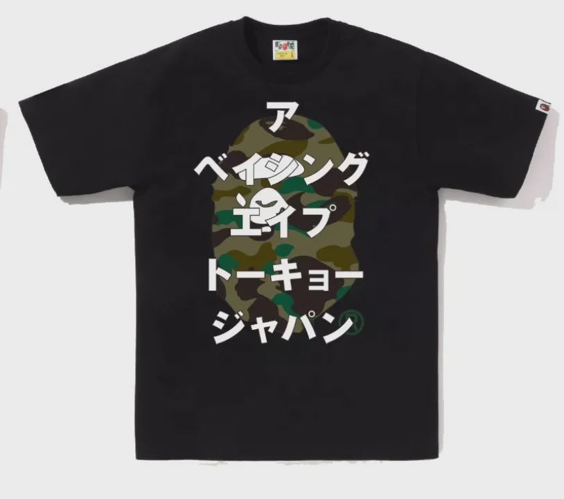 A close-up image of the BAPE 1st Camo Bape Katakana Tee in Black/Green, highlighting the iconic BAPE logo and vibrant camo pattern on the t-shirt