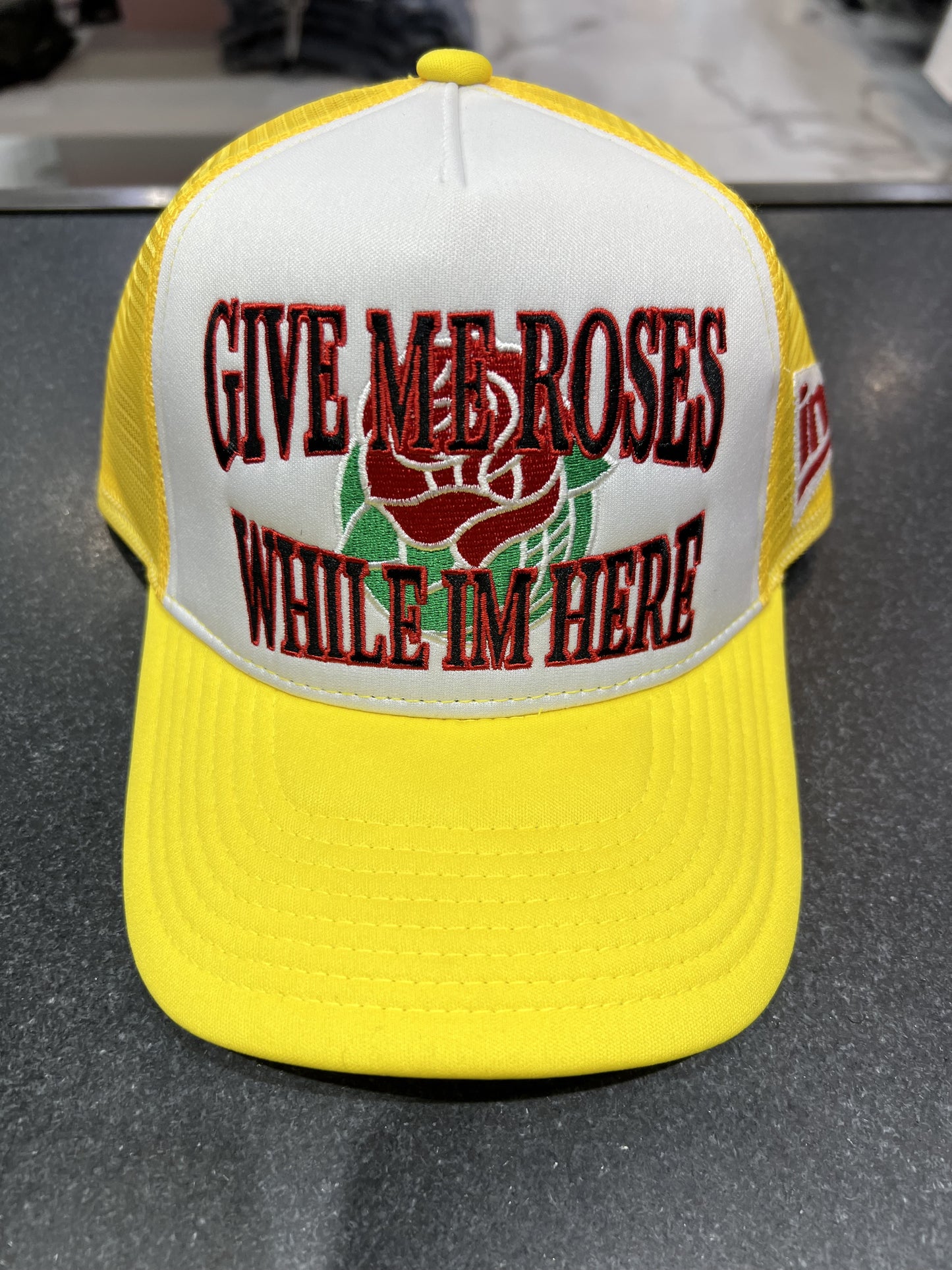 Yellow trucker hat with 'Give Me Roses While I'm Here' design