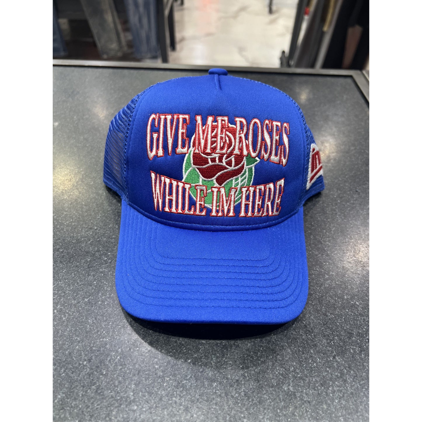 Blue trucker hat with embroidered text 'Give Me Roses While I'm Here'